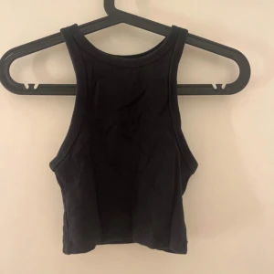 Svart ribbad croppad linnetopp - Säljer en svart croppad linnetopp med ribbad struktur. Toppen har en sportig skärning med breda axelband och hög halsringning. Perfekt att styla till jeans eller kjol.