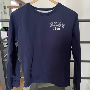 Mörkblå sweatshirt från GANT - Säljer en klassisk mörkblå sweatshirt från GANT med vit broderad logga och årtal '1949' på bröstet. Tröjan har rund halsringning och långa ärmar. Perfekt för en stilren och avslappnad look.