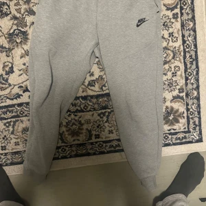 Grå mjukisbyxor från Nike - Säljer ett par grå mjukisbyxor från Nike med logga på vänster höft. Byxorna har resår i midjan och vid bensluten, samt en avslappnad passform. Perfekta för chill eller träning.