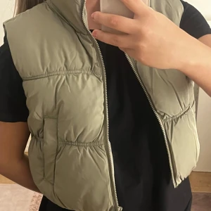 Beige dunväst från H&M - Säljer en beige dunväst från H&M med dragkedja framtill och två fickor. Västen är tillverkad av 100% återvunnen polyester och har en klassisk, vadderad design som passar perfekt till lager-på-lager-stil.