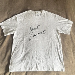 Vit t-shirt från Saint Laurent - Säljer en vit t-shirt från Saint Laurent med svart logga på bröstet. Klassisk passform och rund halsringning. Perfekt för dig som gillar stilrena designer och exklusiva märken. På bilden är 180 cm, 79kg. 