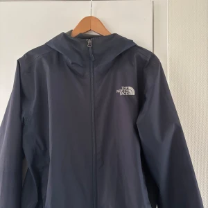 Mörkblå vindjacka från The North Face - Säljer en mörkblå vindjacka från The North Face med huva och dragkedja framtill. Jackan har en stilren design med logga på bröstet och är perfekt för blåsiga dagar. Passar dig som gillar funktionella och snygga ytterplagg.