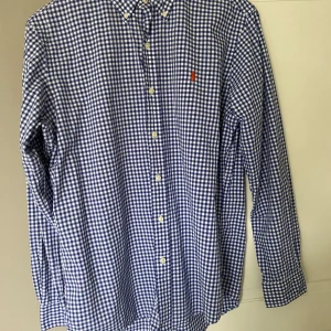 Blårutig skjorta från Ralph Lauren - Klassisk blårutig skjorta från Ralph Lauren med vit knappstängning och broderad logga på bröstet. Skjortan har lång ärm och button-down krage, perfekt för en stilren look. Helt ny, bara att skriva vid funderingar.