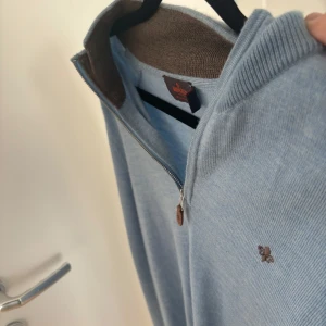 Ljusblå merinoullströja med dragkedja från Morris - Säljer en ljusblå tröja från Morris i 100% merinoull. Tröjan har halv dragkedja vid halsen, ribbade muddar och en liten broderad logga på bröstet. Perfekt för lager-på-lager och snygg till både jeans och chinos.