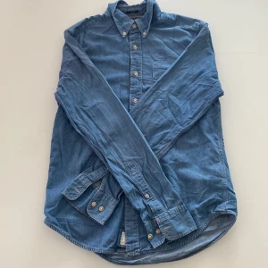 Blå jeansskjorta med knappar - Säljer en klassisk blå jeansskjorta med långa ärmar och knappar hela vägen framtill. Skjortan har button-down krage och är tillverkad i mjukt jeanstyg. Perfekt för en avslappnad stil.