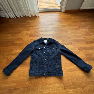 Mörkblå jeansjacka - Säljer en klassisk mörkblå jeansjacka med silverfärgade knappar framtill och på bröstfickorna. Jackan har lång ärm och en rak passform. Perfekt att slänga över en hoodie eller t-shirt för en cool look.