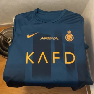 Al Nassr Ronaldo Fotbollströja  - Snygg blå fotbollströja från Al Nassr med guldiga detaljer och tryck. Framsidan har klubbmärke, Nike-logga och sponsortryck, baksidan har namnet Ronaldo och nummer 7 i guld. Perfekt för fans av Ronaldo och Al Nassr.