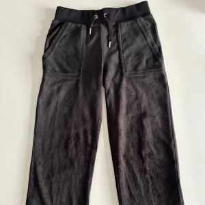 Svarta mjukisbyxor från Juicy Couture - Svarta mjukisbyxor från Juicy Couture med dragsko i midjan och stora fickor fram och bak. Byxorna har en klassisk Juicy Couture-logga på ena fickan och är tillverkade i ett mjukt material som känns lyxigt.