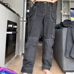 Svarta cargobyxor med vita sömmar - OBS!! ALLA MINA PLAGG GRATIS VID UPPHÄTMNING I RÅCKSTA!!! Säljer ett par svarta cargobyxor/arbetsbyxor med kontrasterande vita sömmar och flera fickor på sidorna. Har de uppvikta på bilden då de är lite för långa för mig som är 169cm. Gjorda i styvt slitstarkt material som liksom de flesta arbetsbyxor blir mjukare för varje användning. Går fint att ha casual också. 