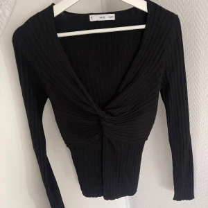 Svart ribbad topp med knut från Mango  - Snygg svart långärmad topp från Mango med ribbad struktur och en knut framtill. Djup v-ringning ger en trendig look. Perfekt att styla med jeans eller kjol.