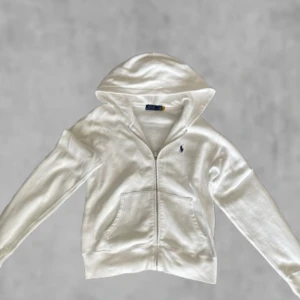 Vit hoodie från Polo Ralph Lauren - Vit hoodie med dragkedja från Polo Ralph Lauren. Klassisk design med huva, två fickor fram och broderad logga på bröstet. Perfekt för en avslappnad och stilren look.