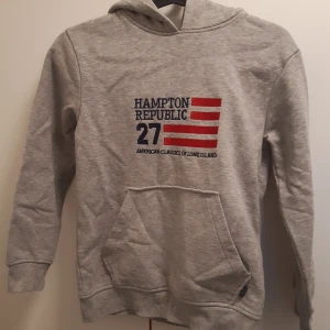 Grå hoodie från Hampton Republic - Ljusgrå hoodie från Hampton Republic med tryck framtill i rött och blått. Tröjan har huva och en stor magficka. Perfekt för en avslappnad stil och enkel att matcha med det mesta.