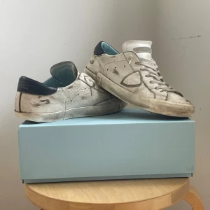 Philippe Model - Säljer ett par vita sneakers från Philippe Model med grå detaljer och svart häl. Skorna har snörning, rund tå och platt sula. Klassisk design med diskret logga på sidan och bekväm innersula. Skorna är väl använda och har många slittningar men fungerar fortfarande fint,  okej skick storlek 42
