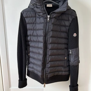 Svart pufferjacka med stickade ärmar från Moncler - Säljer en svart pufferjacka från Moncler med stickade ärmar och huva. Jackan har dragkedja framtill, ribbade muddar och en liten ficka med dragkedja på ena ärmen. Perfekt för kyliga dagar.