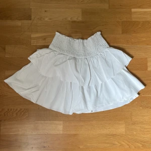 Vit volangkjol  - Säljer en vit kortkjol från SHEIN med bred smock i midjan och dubbla volanger. Kjolen är luftig och har en flowy känsla, perfekt för varma dagar. Enkel att matcha med olika toppar för en fräsch look. Storlek XS