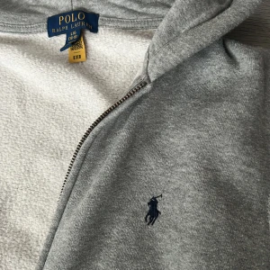 Grå kofta från Polo Ralph Lauren - Säljer en klassisk grå kofta från Polo Ralph Lauren med dragkedja och huva. Tröjan har två fickor framtill och den ikoniska broderade loggan på bröstet. Perfekt för en avslappnad och stilren look.