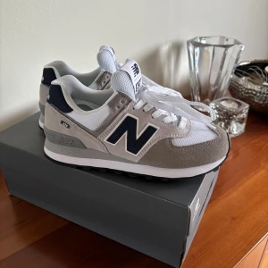 New Balance 574 sneakers - Super snygga New Balance 574 sneakers i grått, vitt och mörkblått. Skorna har klassisk design med mockadetaljer, meshpaneler och snörning. Perfekta för en stilren vardagslook och riktigt bekväma att gå i. Skorna är helt oanvända och perfekta inför sommaren men även nu på våren!😊(Nypris-1345kr!)
