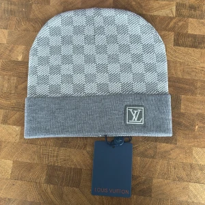 Grå rutig mössa från Louis Vuitton - Snygg grå mössa från Louis Vuitton med diskret rutigt mönster och LV-logga framtill. Mössan har en vikt kant och är perfekt för att hålla värmen med stil.