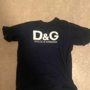  t-shirt från Dolce & Gabbana - Mörkblå t-shirt med vit D&G-logga på bröstet och liten logga i nacken. Klassisk passform och rund halsringning. Perfekt för dig som gillar stilrena märkesplagg.