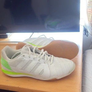 Vita Adidas Sala sneakers med gröna detaljer - Säljer ett par vita Adidas Sala sneakers med gröna detaljer och klassiska ränder på sidorna. Skorna har snörning och platt sula, perfekta för inomhusaktiviteter. Snygg och sportig design med meshpaneler för extra ventilation. Lite små skador från endast 2-3 träningar men inga håll eller större slitningar!