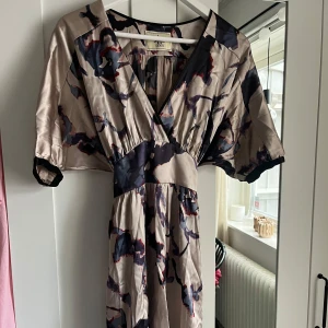Mönstrad omlottklänning från De Moy - Säljer en snygg omlottklänning från De Moy i beige med abstrakt mönster i mörkblått och rött. Klänningen har korta ärmar, v-ringning och knytband i midjan. Perfekt för dig som gillar unika plagg med skön passform. Skriv för fler bilder💞