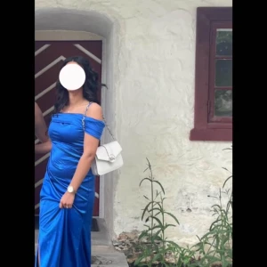 Blå axelbandslös långklänning med slits - Säljer en glansig blå långklänning med offshoulder och smala axelband. Klänningen har en hög slits framtill och figurnära passform, perfekt för bal eller fest. Materialet är satinliknande och ger en lyxig känsla. S/M i storlek 
