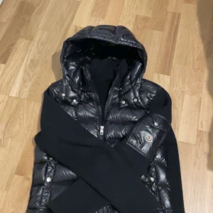 Svart pufferjacka från Moncler - Snygg svart pufferjacka från Moncler med huva och dragkedja framtill. Jackan har långa ärmar i stickat material och klassisk Moncler-logga på ena ärmen. Perfekt för kyliga dagar.