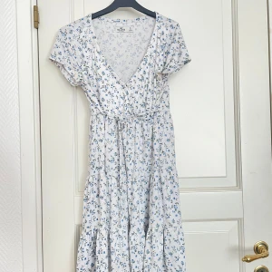 Vit blommig omlottklänning - Söt vit klänning med små blå blommor och omlott-v-ringning. Klänningen har korta ärmar och volangdetaljer nertill, vilket ger en luftig och somrig känsla. Perfekt för varma dagar och midsommarfirande.