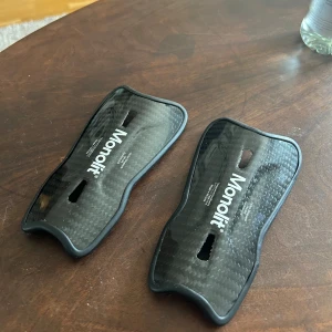 Monolit benskydd (14cm)  - Säljer ett par svarta benskydd från Monolit. De är tillverkade i kolfiber vilket gör de ultralätta och smidiga. Perfekta för fotbollsspelare som vill kunna röra sig obehindrat utan att ens märka av att du har på dig benskydd. 