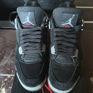 Svarta Nike Air Jordans 4 Retro - Säljer ett par svarta Nike Air Jordans 4 Retro med grå och röda detaljer. Skorna har meshpaneler på sidorna, plastdetaljer vid snörningen och en synlig Air-sula. Ikonisk Jumpman-logga på plösen och undersidan.