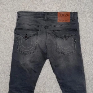 Chief Keef True Religion Jeans - Säljer ett par grå jeans från True Religion med klassiska bakfickor och snygga kontrastsömmar. Jeansen har fem fickor och en skinnpatch med logga bak i midjan. Perfekta för dig som gillar streetwear och vill ha något unikt. Chief Keef liknande Jeans