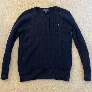 Mörkblå stickad tröja från Polo Ralph Lauren - Säljer en mörkblå stickad tröja från Polo Ralph Lauren med rund halsringning och diskret kabelstickat mönster. Tröjan har lång ärm och en liten grön broderad logga på bröstet. Perfekt för kyligare dagar. Är L i barnstorlek men passar xs i vuxenstorlek