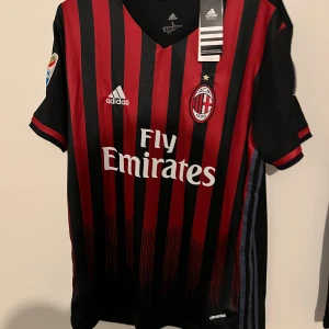 AC Milan hemmatröja Adidas - Säljer en klassisk AC Milan hemmatröja från Adidas med svarta och röda ränder. Tröjan har korta ärmar, broderad klubbmärke, Fly Emirates sponsortryck och Serie A samt Champions League-märken på ärmarna.