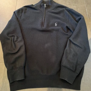 Svart stickad tröja med dragkedja från Polo Ralph Lauren - Säljer en svart stickad tröja från Polo Ralph Lauren med halv dragkedja och broderad logga på bröstet. Tröjan har lång ärm, hög krage och ribbade muddar. Perfekt för en stilren och klassisk look. Storlek s i herr!