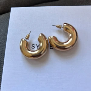 Chunky guldiga örhängen - Säljer ett par chunky örhängen i guldig färg. De är runda och tjocka, med en slät och blank yta. Perfekta för att ge en statement-look till din outfit. Passar dig som gillar trendiga accessoarer.