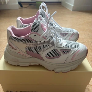 Vita och rosa sneakers från Axel Arigato - Säljer ett par snygga sneakers från Axel Arigato i vitt och ljusrosa med meshdetaljer och kraftig sula. Skorna har snörning och sportig design med rosa detaljer på sula och insida. Perfekta för en trendig vardagslook.