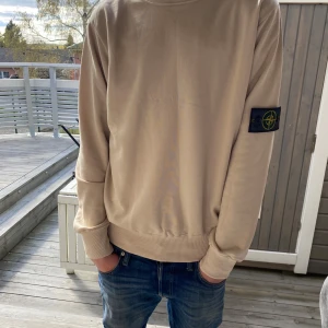 Beige sweatshirt från Stone Island - Säljer en beige sweatshirt från Stone Island med klassisk patch på vänster ärm. Tröjan har rund halsringning, ribbade muddar och är tillverkad i mjukt material. Perfekt för en stilren och avslappnad look.