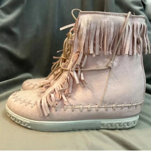 Ljusrosa mockasneakers med fransar - Säljer ett par ljusrosa sneakers i mocka med fransdetaljer längs skaftet och snörning framtill. Skorna har en vit platt sula och dekorativa sömmar runt om. Perfekta för dig som vill sticka ut med en unik stil.