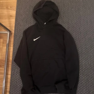 Svart hoodie från Nike - Svart hoodie från Nike med klassisk vit logga på bröstet. Tröjan har huva och en stor magficka framtill. Perfekt för en avslappnad stil och enkel att matcha med det mesta och funkar för träning.