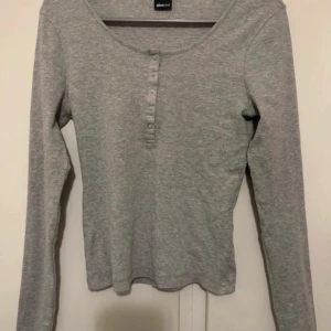 Grå ribbad långärmad topp från Gina Tricot - Grå långärmad topp från Gina Tricot i ribbat material. Rund halsringning och knappdetaljer framtill. Perfekt basplagg som funkar till många olika outfits.