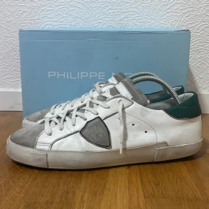 Vita sneakers från Philippe Model med grå och gröna detaljer - Snygga vita sneakers från Philippe Model med grå mockadetaljer på tå och sidor samt grön häl. Skorna har snörning och en platt sula. Perfekta för en stilren och avslappnad look. Skorna är i storlek 42 och dem är i bra skick! Hör av er ifall ni har några frågor! 🙋‍♂️ Nypris runt 3800 kr