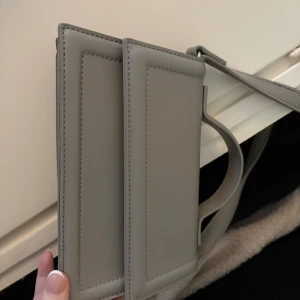 Grå axelväska i syntet - Säljer en stilren grå axelväska med minimalistisk design. Väskan har raka linjer och justerbar axelrem, perfekt för att bära det viktigaste. Enkel och modern look som passar till många outfits.