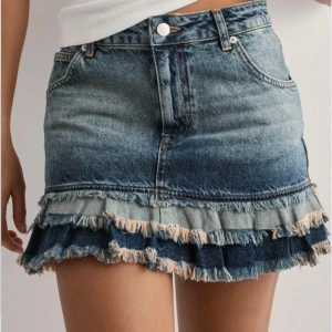 Jeanskjol med volangkant - Säljer en blå jeanskjol från Nelly med volangdetalj och råa kanter längst ner. HELT NY! Prislapp kvar. Ordinarie pris 599kr