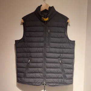 Mörkgrå dunväst från Parajumpers - Säljer en mörkgrå dunväst från Parajumpers i modellen Super Lightweight. Västen har dragkedja framtill, två fickor med dragkedja och en hög krage med gul detalj. Perfekt för lager-på-lager under kyliga dagar.