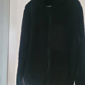 Svart teddyjacka från Solid - Mysig svart teddyjacka från Solid med huva. Jackan har lång ärm och är tillverkad i ett mjukt teddy-material som passar perfekt för kyligare dagar. Enkel och stilren design som funkar till många outfits.