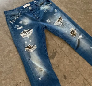 Blå slitna jeans från Replay Broken Edge - Säljer ett par blå jeans från Replay Broken Edge med slitningar och hål framtill. Jeansen har klassisk femficksdesign, raka ben och normal passform. Perfekta för dig som gillar en tuff och avslappnad stil.