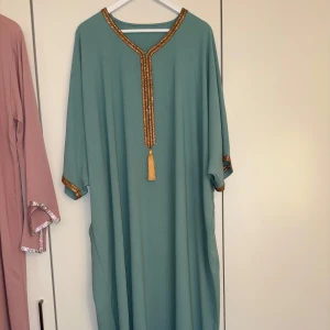 Ljusgrön kaftanklänning med guldiga detaljer - Säljer en ljusgrön lång kaftanklänning med vackra guldiga broderier längs halsringningen och ärmsluten. Klänningen har en lös passform och dekorativ tofs framtill. Perfekt för dig som gillar en elegant och bekväm stil. (Marockansk klänning) 
