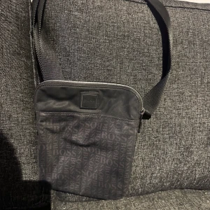 Svart axelväska från BOSS - Snygg svart axelväska från BOSS med logotypmönster över hela väskan. Väskan har justerbar axelrem och dragkedja upptill. Perfekt för att bära med sig det viktigaste i vardagen.