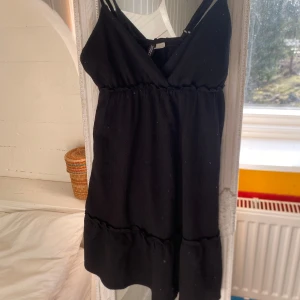 Svart kortklänning från H&M - Säljer en svart kortklänning från Divided H&M med v-ringning och smala axelband. Klänningen har en volang nertill och är tillverkad i ett lätt, strukturerat tyg. Perfekt för varma dagar eller att styla med en jacka.