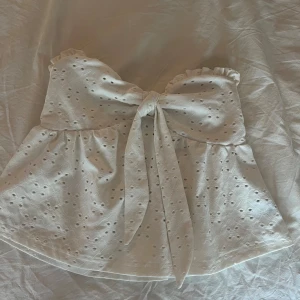 Vit broderad tubtopp med rosett - Superfin vit tubtopp med broderade hål och stor rosett framtill. Toppen har volangkant upptill och är axelbandslös, perfekt för sommardagar. Söt och luftig modell som passar till både jeans och kjol.